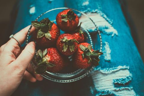 Hand holding bowl of ripe strawberries on ripped denim lap with ring, moody rustic snack