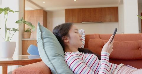 Asian woman relaxing on sofa using smartphone in contemporary living room