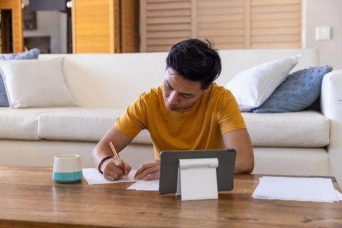Focused Asian man in twenties writing papers at table with tablet and mug in casual home