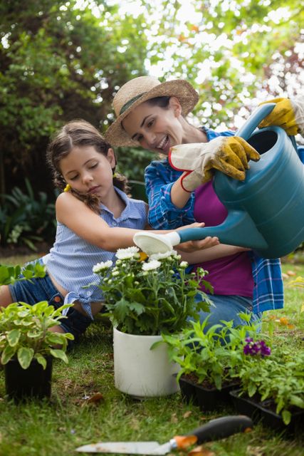 Happy mother with daughter watering potted plants while sitting on field in backyard