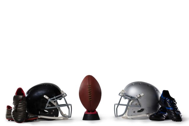 This image shows an American football placed on a tee, flanked by two helmets and pairs of sports shoes on either side, all set against a white background. Ideal for use in sports-related articles, promotional materials for football events, team merchandise advertisements, and athletic gear catalogs.