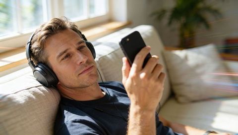 Man relaxing on sofa listening to music with headphones and smartphone in sunlit room