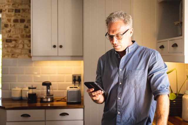 Mature man wearing glasses and using smartphone in a modern kitchen. Ideal for themes related to technology use, home lifestyle, communication, and domestic life. Suitable for articles, blogs, and advertisements focusing on middle-aged individuals, home settings, and modern living.