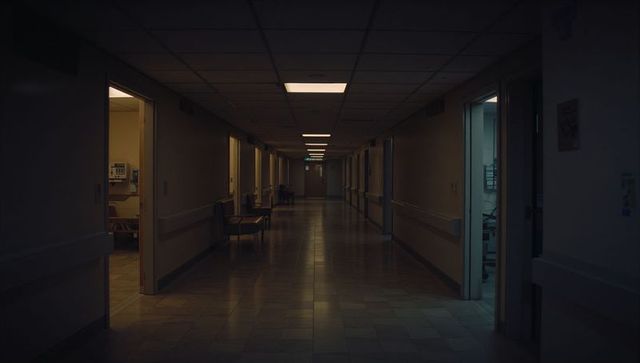 Dim hospital corridor leading to exit, recessed lights, empty patient rooms and handrails