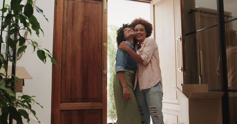 Women embracing while entering new home with moving boxes in sunlit foyer