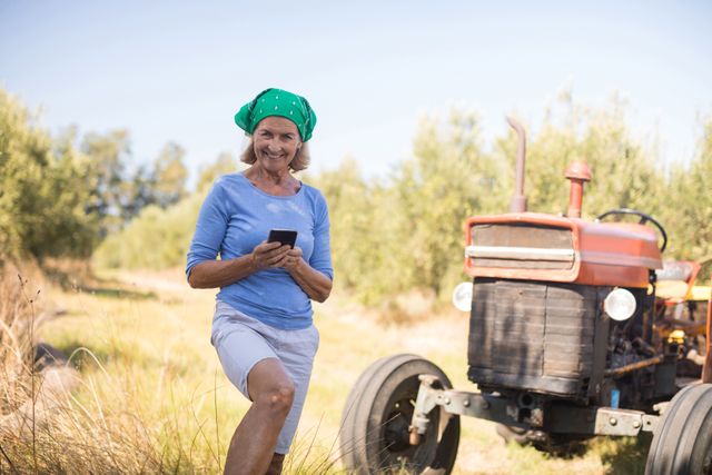Middle-aged woman standing in olive farm using mobile phone, smiling. Tractor in background. Ideal for themes related to agriculture, rural lifestyle, technology in farming, and outdoor activities.