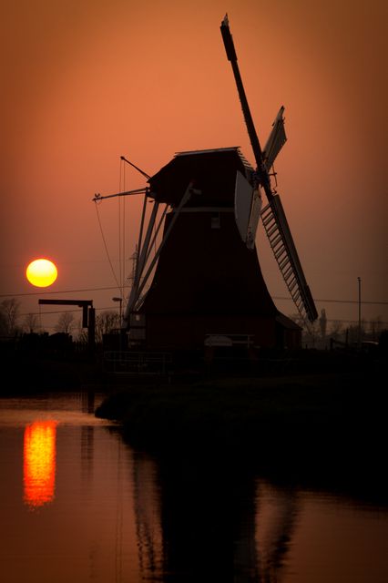 A beautiful silhouette of a traditional Dutch windmill standing by the water during sunset. The vibrant orange sun and its reflection create a serene and peaceful atmosphere, enhanced by the countryside surroundings. Perfect for travel-themed content, nature background, or promoting rural tourism.