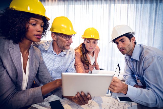 Group of architects wearing hard hats collaborating on a project using a digital tablet in an office. Ideal for illustrating teamwork, professional collaboration, construction planning, and the use of technology in architecture and engineering projects.