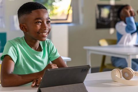 African american boy using tablet and smiling in bright classroom with headphones nearby