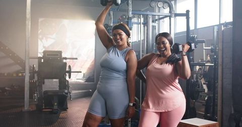 Smiling Black women lifting dumbbells and posing in pastel activewear during gym workout