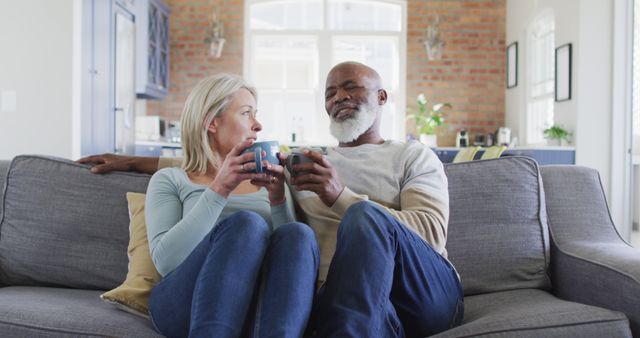 Senior couple sitting on sofa enjoying coffee in cozy modern living room. They appear relaxed and content while sharing a moment of togetherness. Ideal for depicting senior lifestyle, retirement happiness, home comfort, or family bonding in marketing materials, ads, or articles.