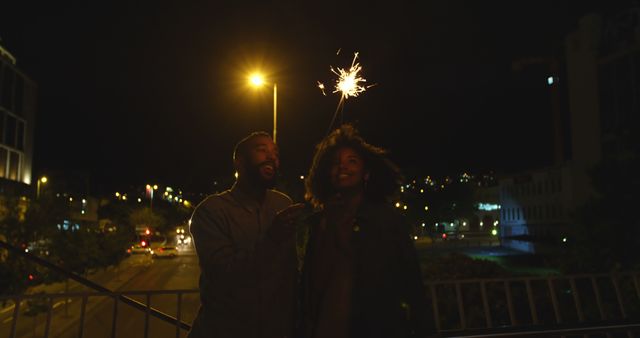 Romantic diverse couple standing and holding sparklers in city street at night. City living, romance, love, relationship, free time and lifestyle, unaltered.