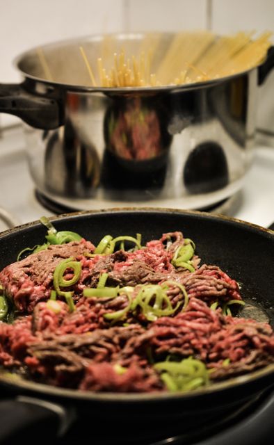 Raw ground beef with slices of green onions in frying pan is being prepared, situated near a pot of cooking spaghetti on a stove. Perfect for blogs and articles about cooking, homemade recipes, pasta dishes, or culinary tips.