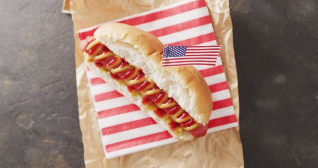 Hot dog topped with ketchup and mustard on brown paper and American flag-themed napkin. Great for use in barbecue, summer, holiday, Independence Day, and picnic promotional materials.