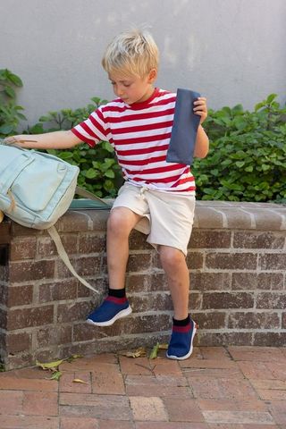 Blond schoolboy sitting on brick wall reaching for mint backpack while holding pencil case