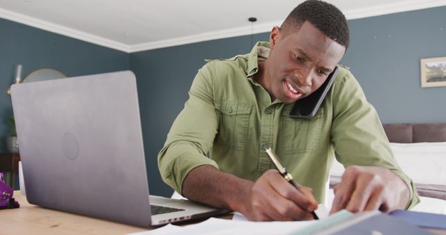Image of african american man using laptop, talking smartphone and writing notes, working in bedroom. Communication, working from home, domestic life, and inclusivity concept.