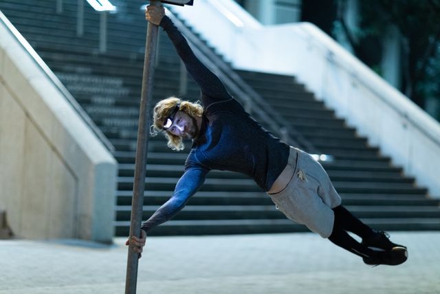 Front view of a fit Caucasian man with long blonde hair wearing sportswear exercising outdoors in the city at the evening, doing a human flag position on the street sign, wearing head light.