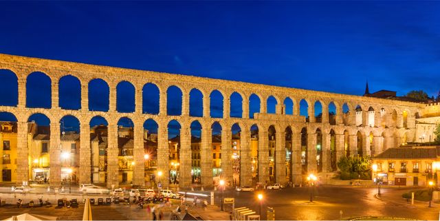 Ancient Roman Aqueduct spanning cityscape, illuminated against deep blue twilight sky in Segovia, Spain. Historic arches showcasing Roman engineering prowess. Perfect for travel blogs, historical documentaries, architectural studies, tourism promotion.