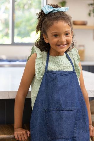 Smiling girl wearing denim apron and polka-dot bow leaning on bright kitchen island