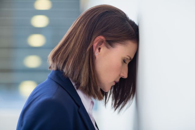 Businesswoman experiencing stress and frustration, leaning her head against a wall in an office environment. Suitable for illustrating workplace stress, mental health issues, corporate burnout, and professional challenges. Can be used in articles, blogs, and presentations related to business, mental health, and work-life balance.
