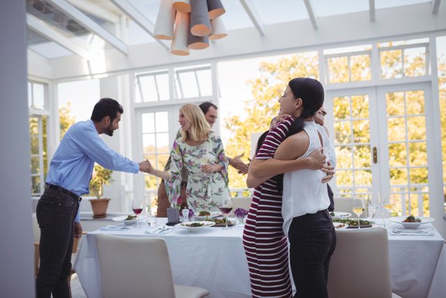 Friends are embracing and greeting each other during a lunch gathering in a bright, modern restaurant. The atmosphere is cheerful and casual, with a table set for dining. This image can be used for themes related to friendship, social gatherings, celebrations, and dining experiences.