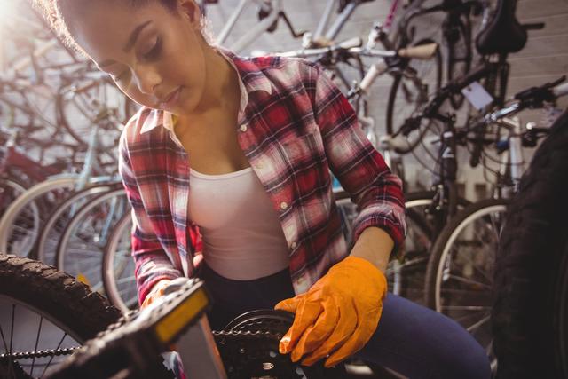 Young woman wearing plaid shirt and gloves repairing a bicycle cog in a bike shop. She is focused on the task, with several other bicycles in the background. Useful for themes related to bicycle repair, women in mechanics, workshops, and cycling maintenance.