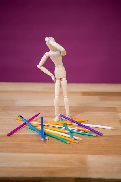 Wooden figurine standing on a wooden table, looking at scattered colorful pencils with a purple background. Ideal for concepts related to creativity, education, artistic inspiration, and design. Suitable for use in educational materials, art-related content, and creative thinking promotions.