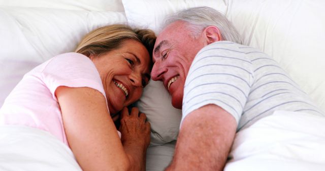 Elderly couple lying in bed, smiling and enjoying each other's company. Perfect for use in themes related to senior love, intimate relationships, retirement living, and mental well-being.