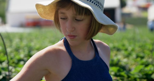 Young girl wearing a large sun hat enjoying the outdoors in a farm or garden setting. Perfect for themes involving childhood, nature, summer activities, gardening, and peaceful rural life.