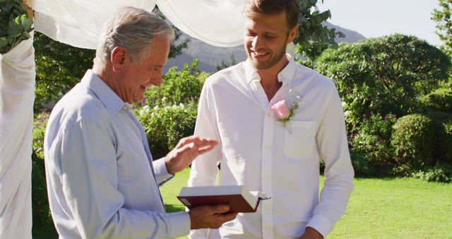 An elderly man in a light blue shirt is handing a book to a smiling groom in a white shirt at an outdoor wedding. The event takes place with lush greenery and mountains in the background under a white arch. Ideal for content about weddings, family traditions, intergenerational bonding, and special moments in nature.