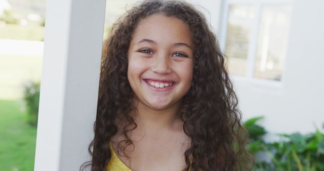 Young girl with long, curly hair smiling joyfully while standing outside. Ideal for advertisements, educational materials, family-oriented projects, blogs, and websites promoting positivity and children's activities.