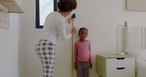 Mother measuring daughter's height with tape in cozy bedroom