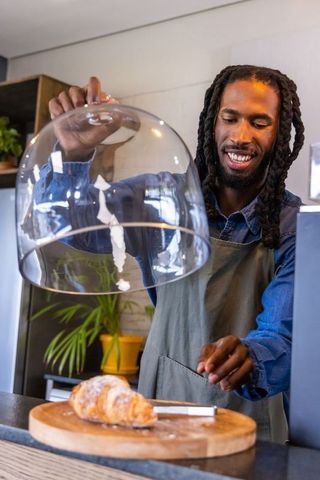 African-american barista lifting glass dome revealing croissant on wooden board, smiling