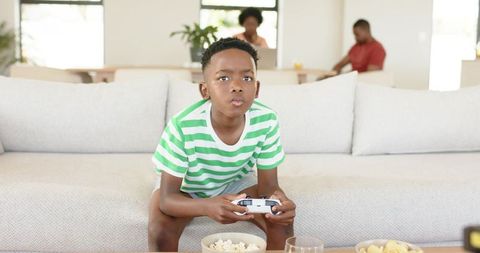 Young african american boy playing video games on living room couch with snacks