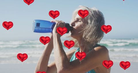 Elderly Woman Hydrating on Beach with Red Heart Balloons