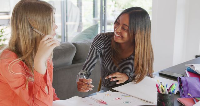 Two young women are collaborating on a creative project surrounded by colorful pens and papers in a well-lit home environment. This can be used to represent teamwork, brainstorming sessions, studying together, casual home learning, and friendship moments.