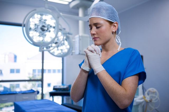 This image depicts a female nurse in surgical attire, standing in an operating room with her hands clasped in prayer. The surgical lights and medical equipment in the background suggest a hospital environment. This image can be used in articles or materials related to healthcare, medical professions, the importance of mental preparation in medical settings, or the role of spirituality and prayer in stressful professions.