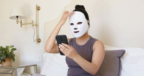 Woman using led facial mask and smartphone on bed for at-home skincare routine