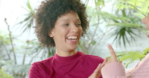 Two diverse women laughing and chatting in bright plant-filled sunroom, candid lifestyle