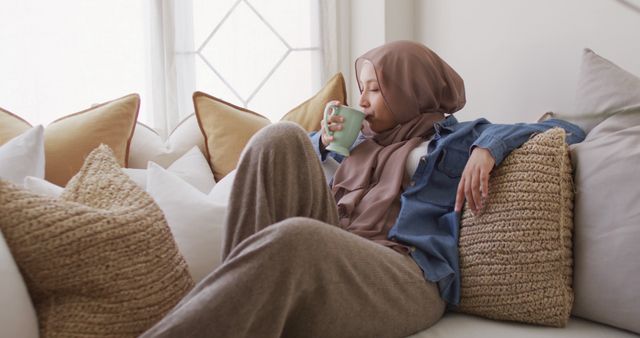 Image of smiling biracial woman in hijab drinking coffee relaxing on sofa at home. Happiness, relaxation, inclusivity and domestic life.