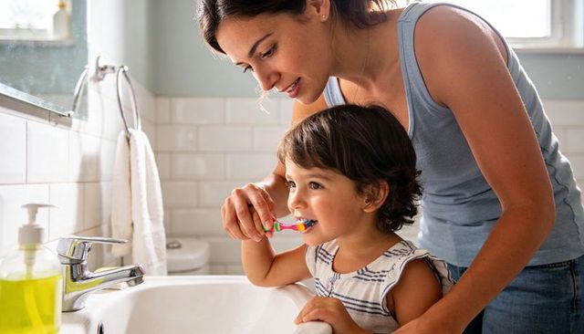 Mother guiding toddler brushing teeth at bathroom sink morning oral care routine