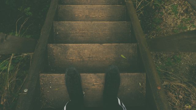 A downward view of a person standing on old wooden stairs outdoors. Useful for themes like exploration, nature walks, hiking, adventure, rustic living, or mindfulness.