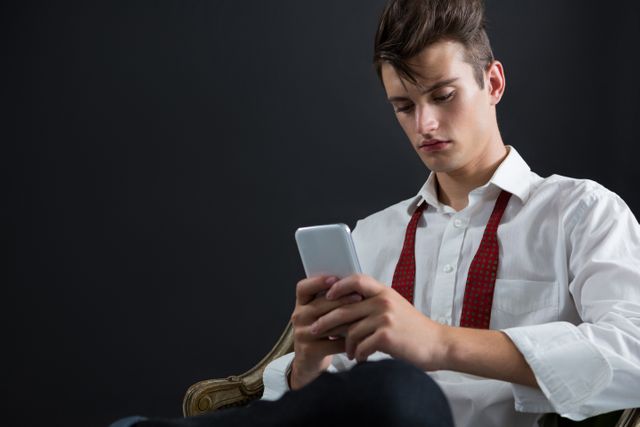 Young androgynous man in formal attire sitting on chair, using mobile phone against black background. Ideal for themes related to modern communication, technology, fashion, and youth culture. Suitable for use in advertisements, blogs, and articles focusing on mobile technology, fashion trends, and lifestyle.