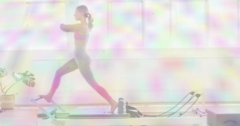 Woman lunging on pilates reformer in bright studio wearing pink bra and grey leggings