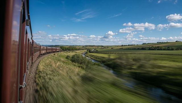 Maroon train curving through lush countryside with stream and blue sky