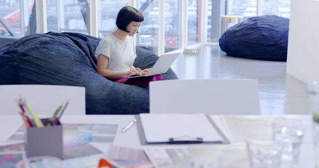 Woman working on her laptop while sitting on a bean bag chair in a modern, creative office. Desk with markers, papers, and other office supplies visible in foreground. Ideal for illustrating concepts such as remote work, casual work environments, coworking spaces, and modern workplace design.