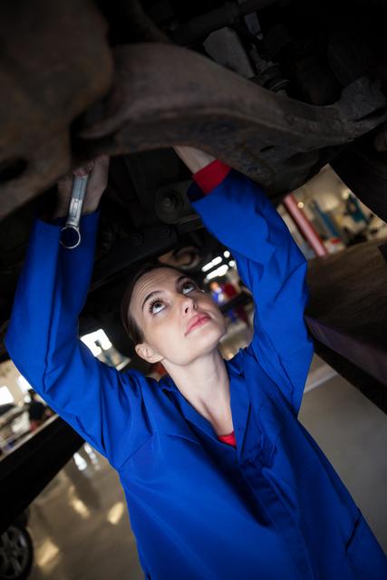 Close-up view of a female mechanic inspecting a car undercarriage in a workshop. She wears blue coveralls, concentrating on her task. This image can be used in articles or advertisements related to automotive services, gender diversity in trades, or professional mechanics.