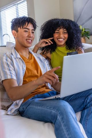Diverse couple relaxing on couch using laptop and smartphone for online shopping at home