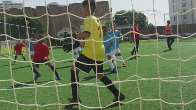 Children enthusiastically play a soccer game on a green artificial turf football field under sunny skies. Capturing teamwork and athleticism, this scene is perfect for sport-related articles, advertisements for youth leagues, or physical activity promotions.