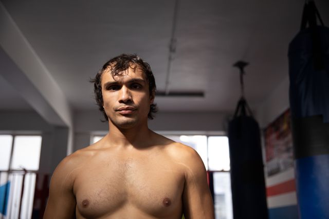 Portrait of a young biracial male boxer with short dark hair, shirtless, in a boxing gym looking straight to the camera.